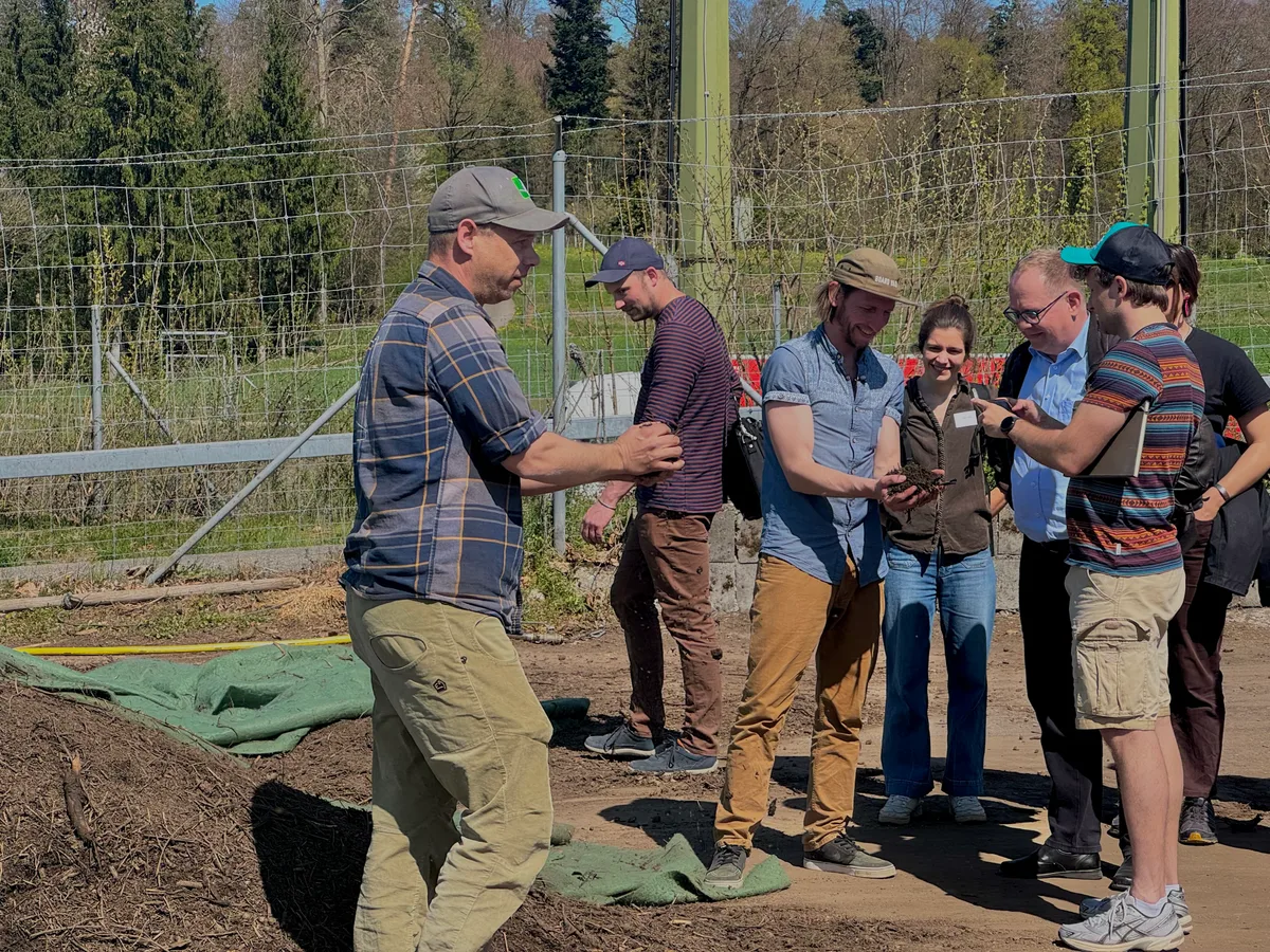 Kompost soll krümelig sein und nach Waldboden riechen: Michael Kunz von der Kunz Baumschulen AG in Uster erläutert den Anwesenden den Prozess des Kompostierens. Im Hintergrund Jojo Casanova von Kompotoi. Zwei Männer halten Kompost in ihren Händen und halten ihn einer Gruppe Menschen zum Beschnuppern hin.